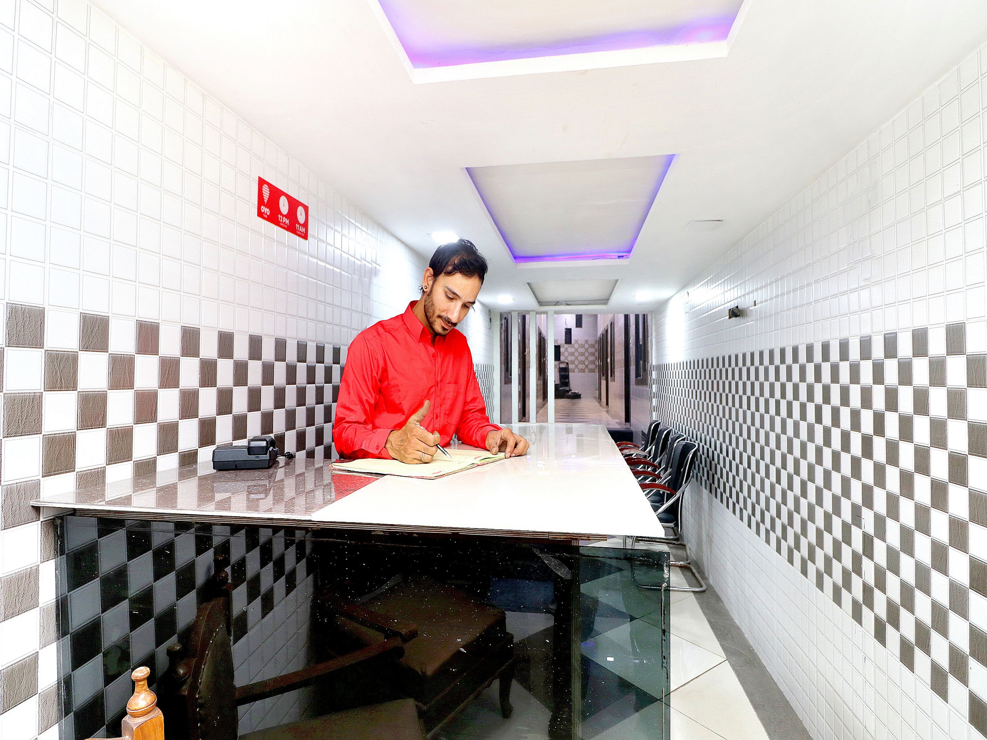 Una persona sentada a una mesa en un pasillo con paredes a cuadros, un techo con una lámpara y un letrero rojo en la pared.