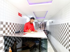 Una persona sentada a una mesa en un pasillo con paredes a cuadros, un techo con una lámpara y un letrero rojo en la pared.