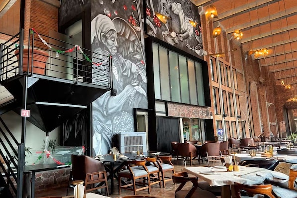 A restaurant with high ceilings, exposed brick walls, and a mural of a chef. Tables are set with chairs, and there is a balcony with railings. - Esplendor by Wyndham San Luis Potosi Monarca (San Luis Potosi)