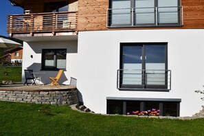 A modern two-story house with a balcony, a stone wall, and a small garden with flowers. - New 2-room holiday apartment in Murnauer Moos (Schwaigen)
