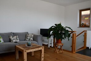 A living room with a grey sofa, a wooden coffee table, a large potted plant, a television, and a white side table. - New 2-room holiday apartment in Murnauer Moos (Schwaigen)