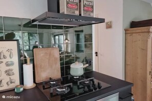 A kitchen with a gas stove, a pot on the burner, a roll of paper towels, and a wooden cutting board. - Oudveluwe - vakantiehuisje - bos (Gelderland)