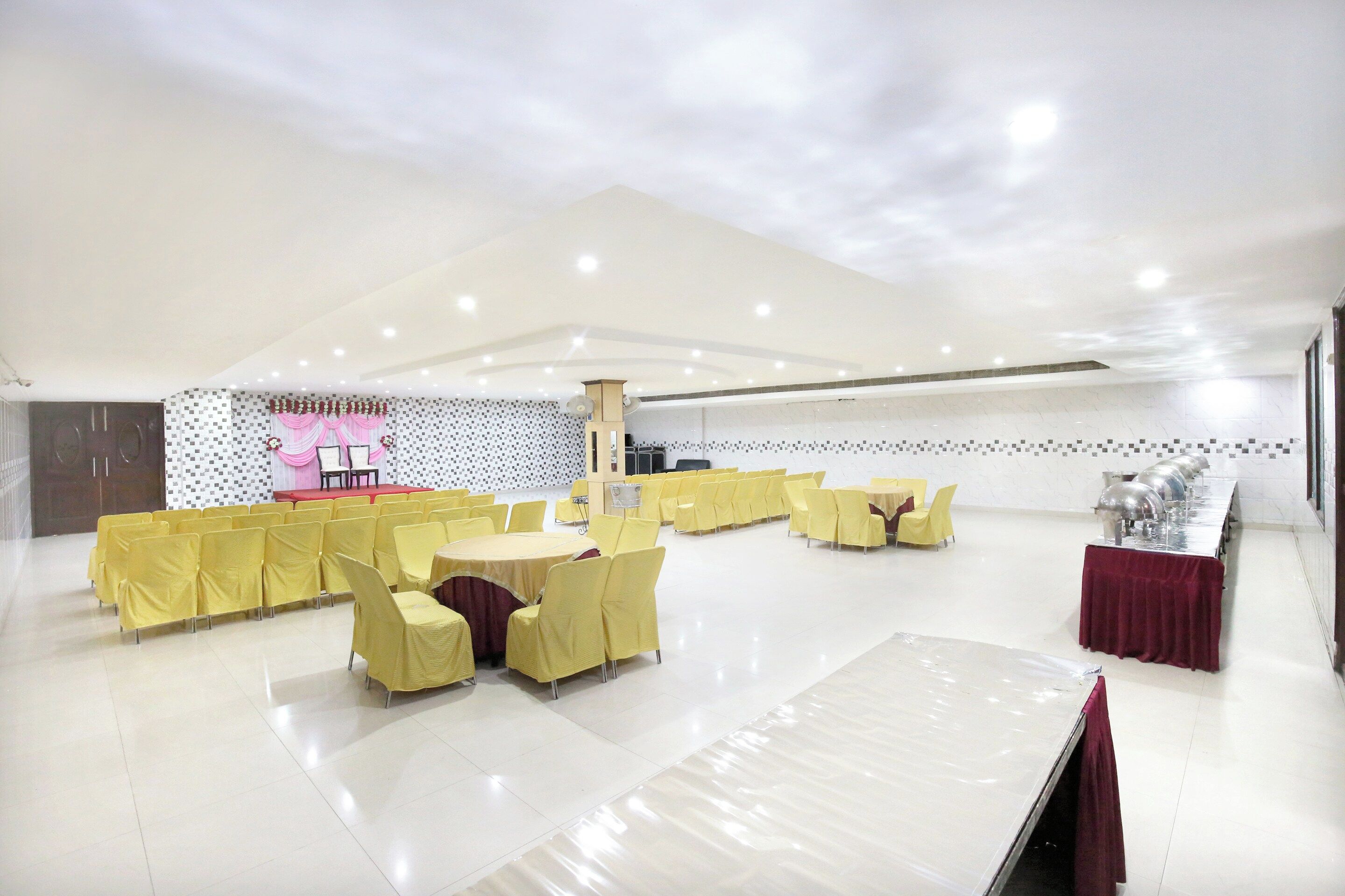 Un salón amplio con hileras de sillas y mesas amarillas, un escenario con cortinas rosas y una pared de azulejos con un diseño geométrico.