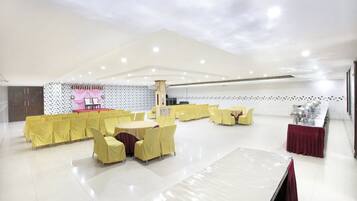 Un salón amplio con hileras de sillas y mesas amarillas, un escenario con cortinas rosas y una pared de azulejos con un diseño geométrico.