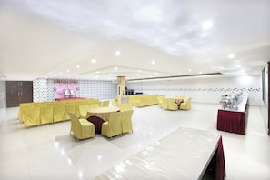 Un salón amplio con hileras de sillas y mesas amarillas, un escenario con cortinas rosas y una pared de azulejos con un diseño geométrico.