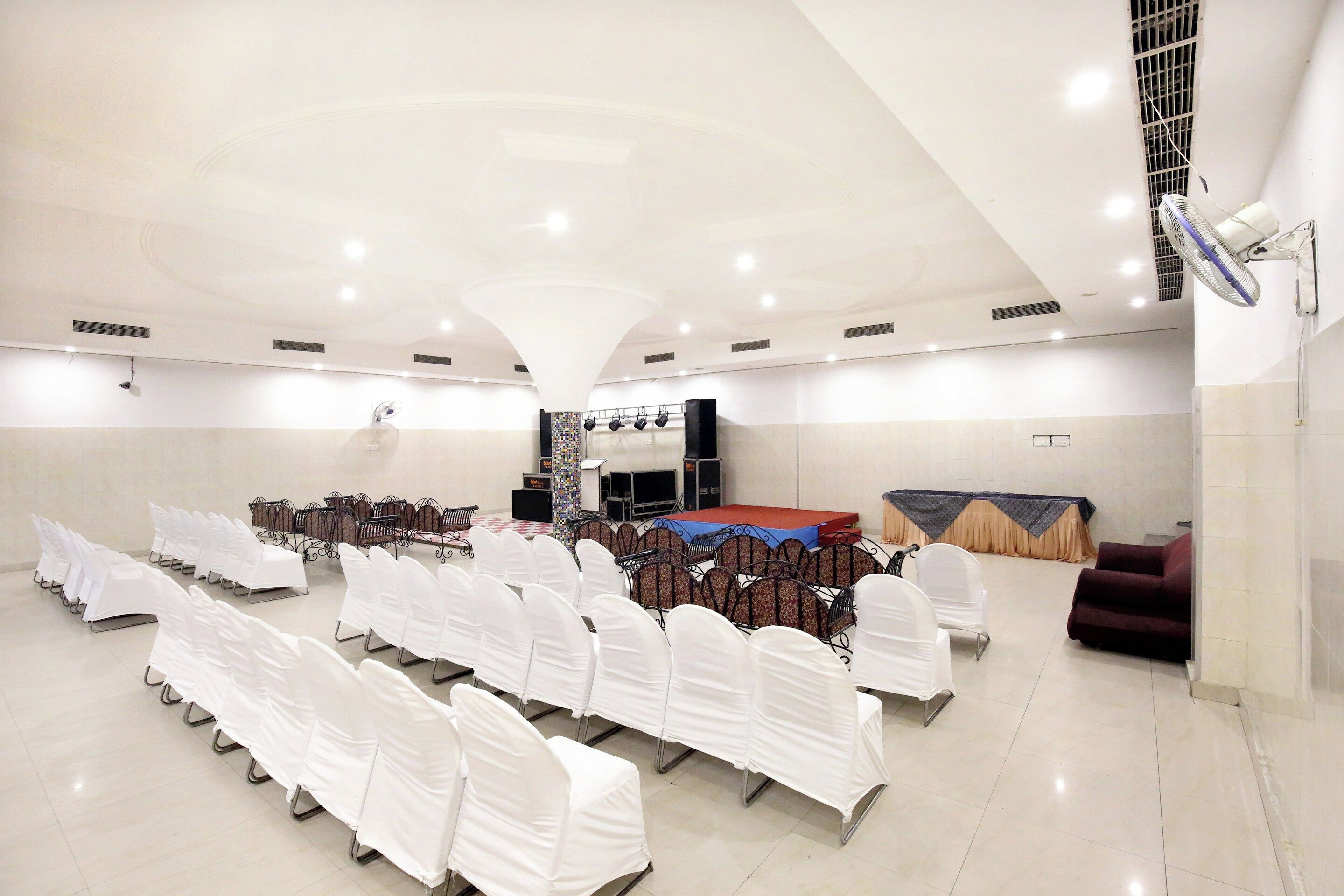 Un salón amplio con filas de sillas blancas, un escenario con plataforma roja y un ventilador de techo.