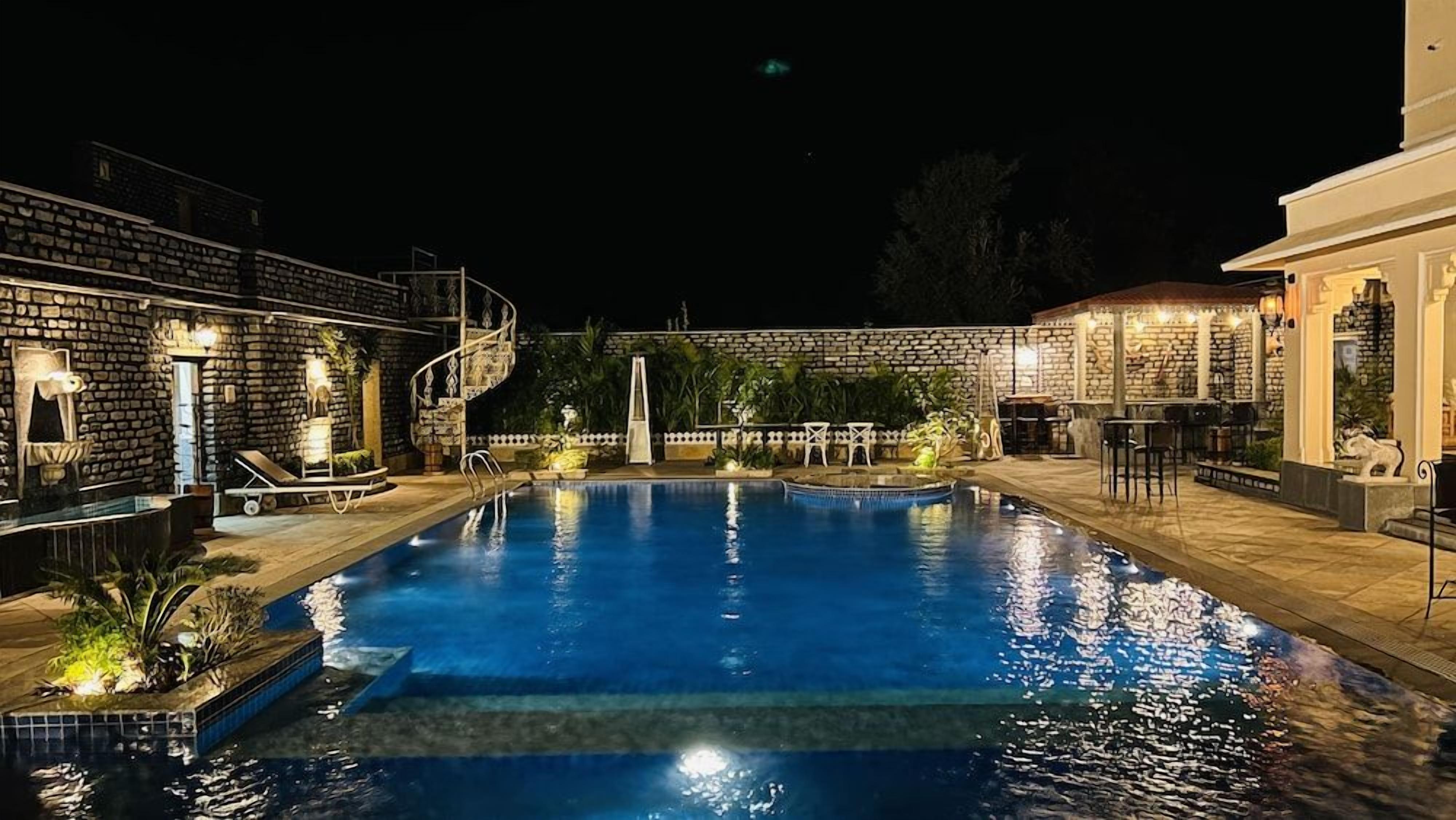 A nighttime swimming pool area with a stone wall, lounge chairs, and a staircase leading to an upper level.