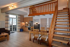 A spacious living area with a wooden staircase, a dining table with chairs, and a kitchen in the background. - Holiday apartment with 1 bedroom (Bodenmais)