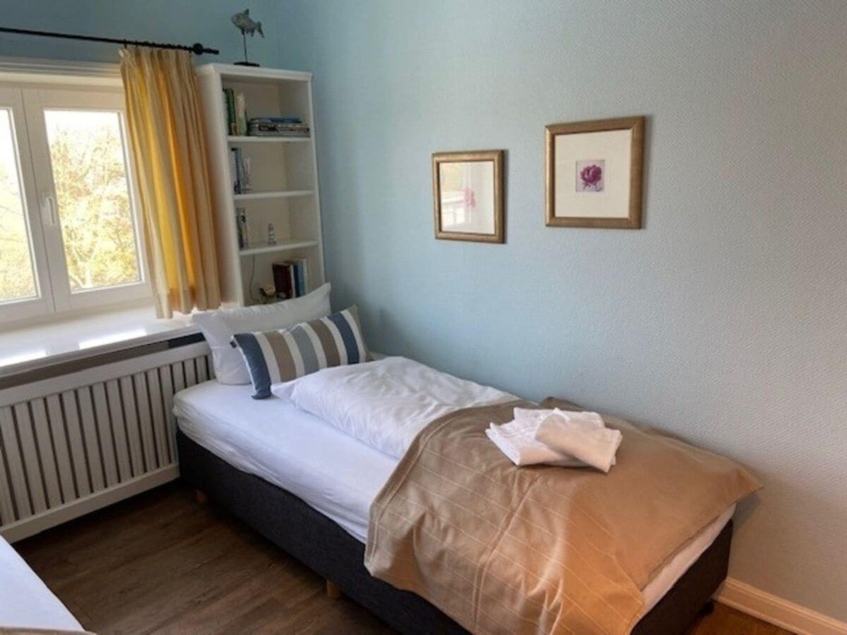 Un dormitorio con una cama individual, una ventana con cortinas, un radiador, una estantería y dos cuadros en la pared.