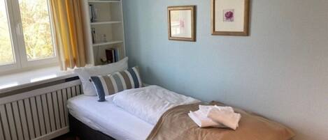 Un dormitorio con una cama individual, una ventana con cortinas, un radiador, una estantería y dos cuadros en la pared.