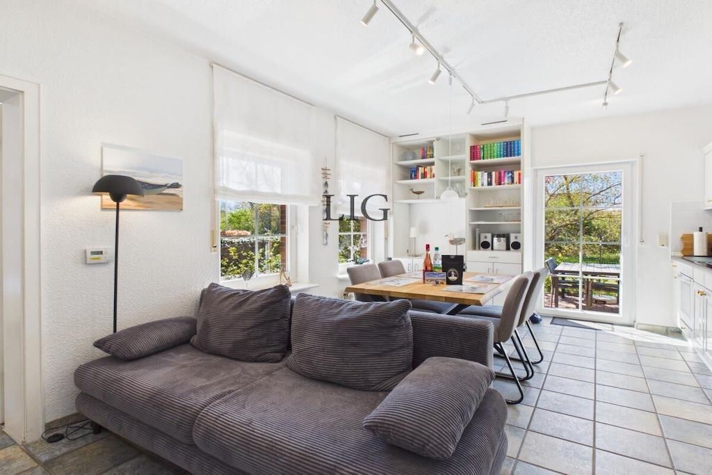 A modern living room with a grey sofa, a dining table with chairs, a bookshelf, and a large window.
