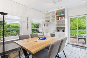 A modern dining area with a wooden table, chairs, a bookshelf, and a view of greenery. - Holiday apartment Seal Bank (Langeoog)