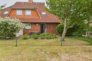 A two-story house with a red-tiled roof, a visible chimney, and a fenced yard with a tree. - Holiday apartment Seal Bank (Langeoog)
