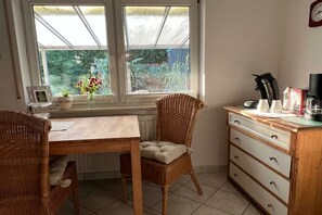 Eine Küche mit Esstisch, Korbstühlen, einem Holzkasten und einem Fenster mit Blick auf Grünflächen.