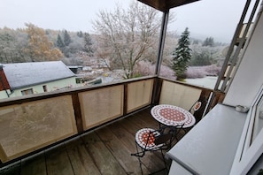 A covered balcony with a wooden floor, two small round tables with chairs, and a view of a snowy landscape with trees and buildings. - Holiday apartment with 2 bedrooms (Kurort Oberwiesenthal)