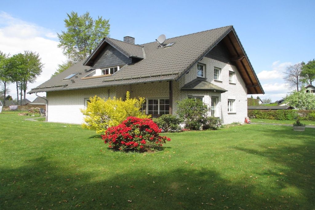 Ein zweigeschossiges Haus mit einem geneigten Dach, einer Satellitenschüssel und einem gepflegten Rasen mit einem roten Blätterbusch.