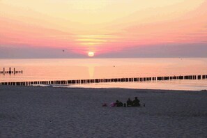 En strandscene med solnedgang, en moler og mennesker siddende i sandet.