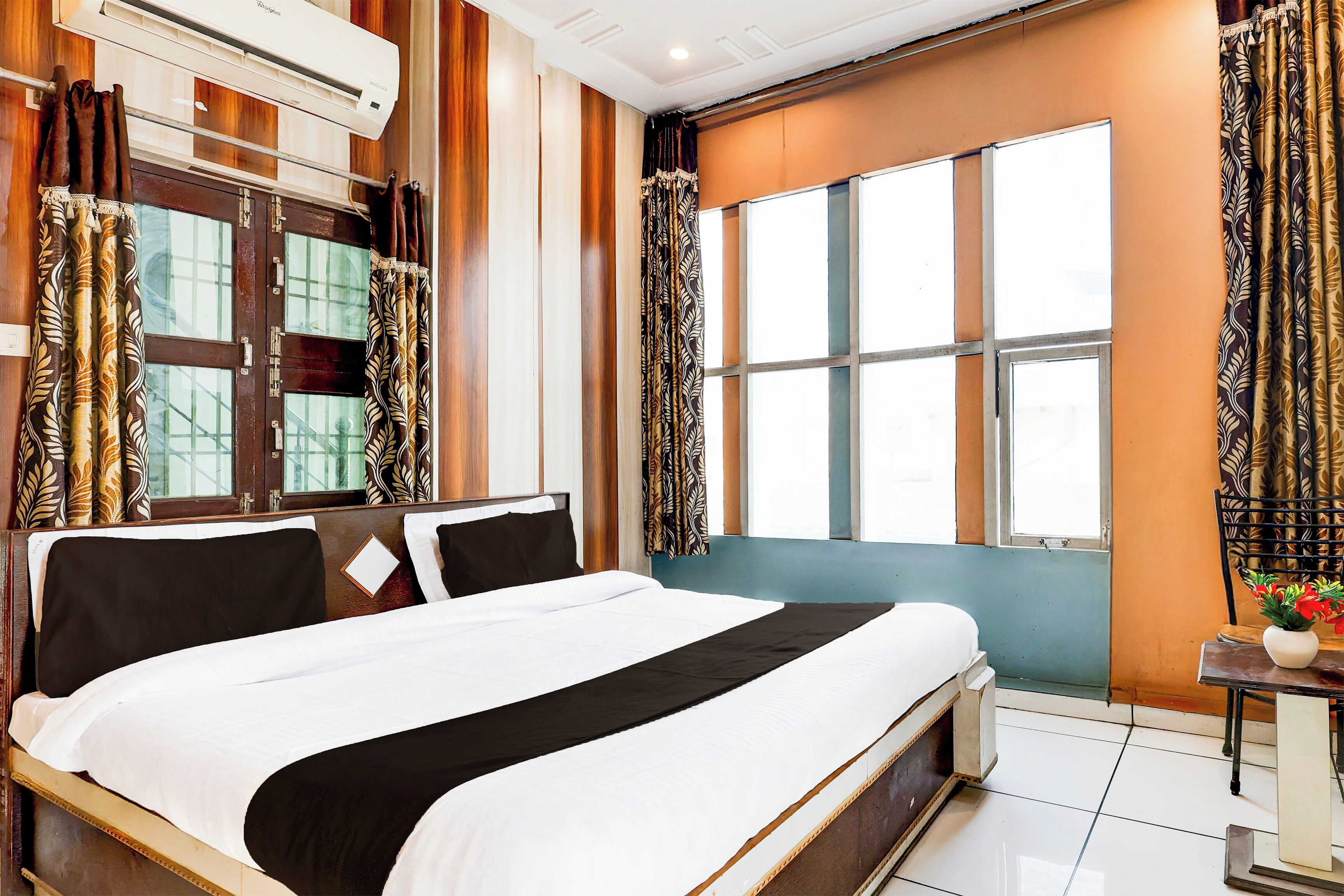 Habitación de hotel con una cama grande, una ventana con cortinas, una mesita de noche con un florero y un equipo de aire acondicionado.
