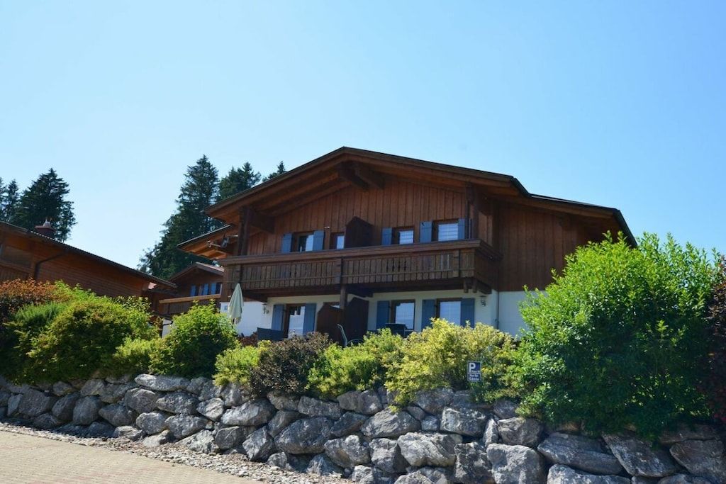 A two-story wooden building with balconies, surrounded by greenery and a stone wall.