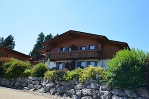 A two-story wooden building with balconies, surrounded by greenery and a stone wall. - Holiday home with 2 bedrooms (Lechbruck am See)