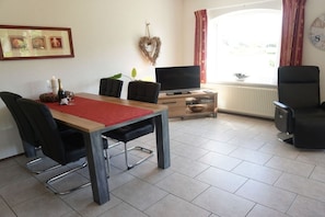 Un salon meublé comprenant une table à manger, des chaises, un téléviseur à écran plat et une fenêtre avec des rideaux.