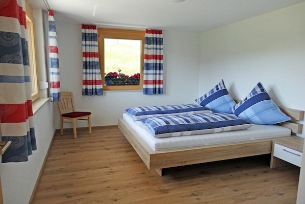 Ein Schlafzimmer mit einem hölzernen Bett, einem Fenster mit Vorhängen, einem Stuhl und einem kleinen Nachttisch.