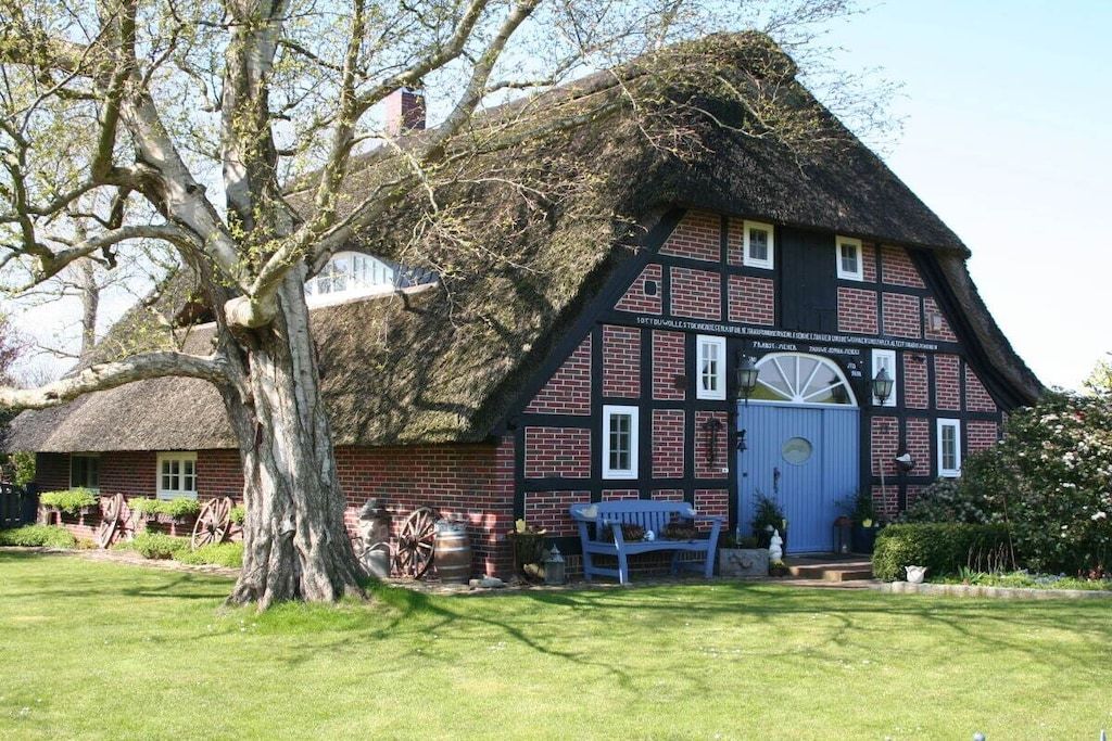 Une maison traditionnelle à colombages avec un toit de chaume, un grand arbre au premier plan et un banc à l’extérieur.