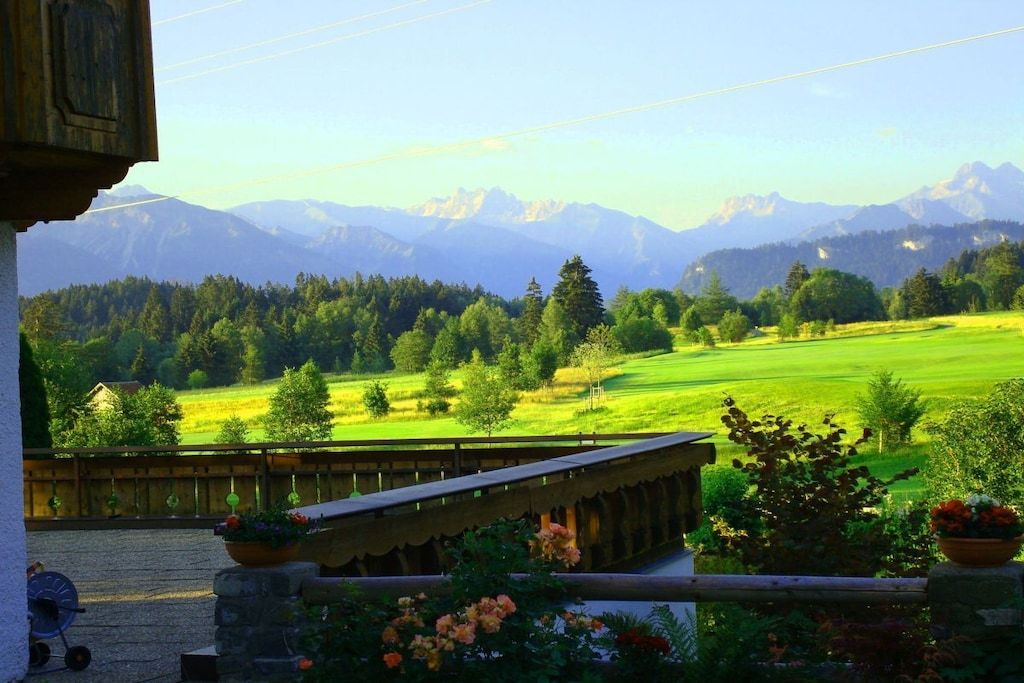 A view from a balcony with a wooden railing, overlooking a green field and mountains in the distance.