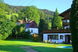 A rural setting with a white building with a red roof, a wooden structure with a balcony, and a clear view of a forested hill in the background. - Holiday apartment with 2 bedrooms (Ofterschwang)