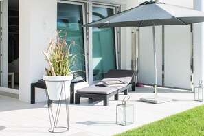 A modern outdoor seating area with a lounge chair and umbrella, a potted plant, and a clear glass door leading to a building. - Timmare III "Porthole" (Timmendorfer Strand)