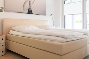 Una cama con ropa de cama blanca, una lámpara de noche y una ventana con vistas.