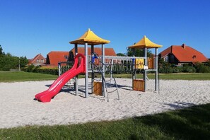 A playground with a red slide and yellow shelters, set on sandy ground with houses in the background. - Holiday home in Neßmersiel with Wi-Fi (Neßmersiel)