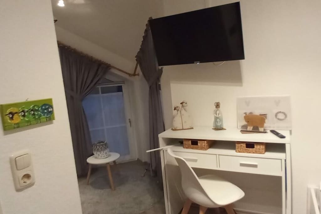 Ein kleines Zimmer mit einem weißen Schreibtisch, einem weißen Stuhl, einem Fernseher über dem Schreibtisch und einem kleinen Tisch mit einer Glasvase.