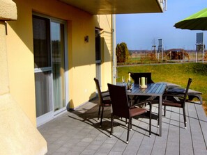 Um pátio externo com mesa e cadeiras, uma porta de vidro e vista para a paisagem.