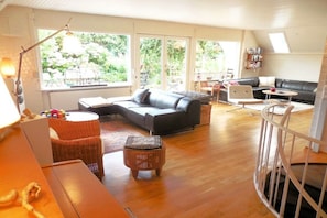 A spacious living room with a sofa, a coffee table, and a dining area with a table and chairs. There is a window providing a view of the outdoors. - Holiday home with 3 bedrooms (Westerholz)