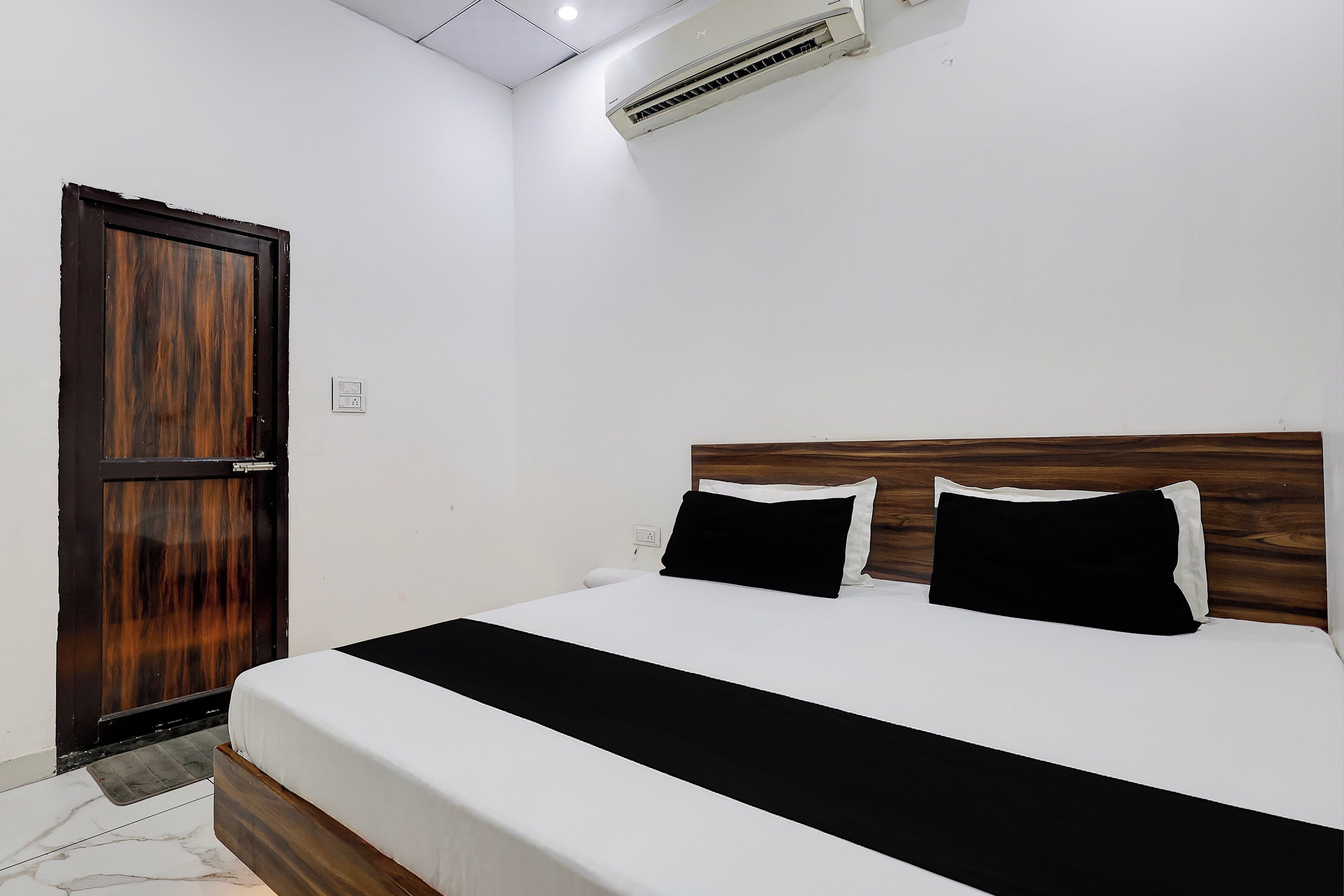 A bedroom with a bed, a wooden door, an air conditioning unit, and a white wall.