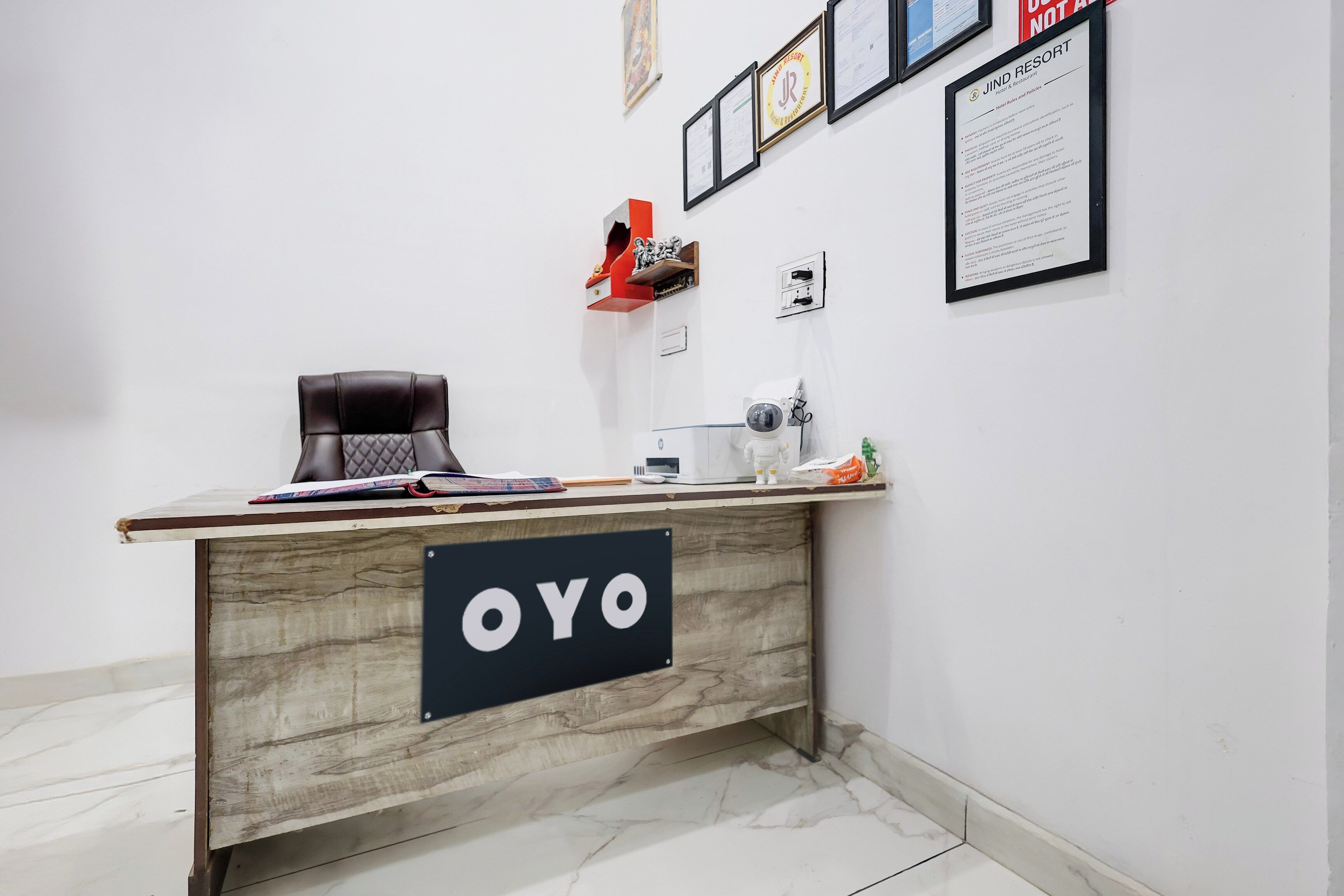 A reception desk with a sign reading 'OYO,' a white wall with framed documents, and a small statue on a shelf.