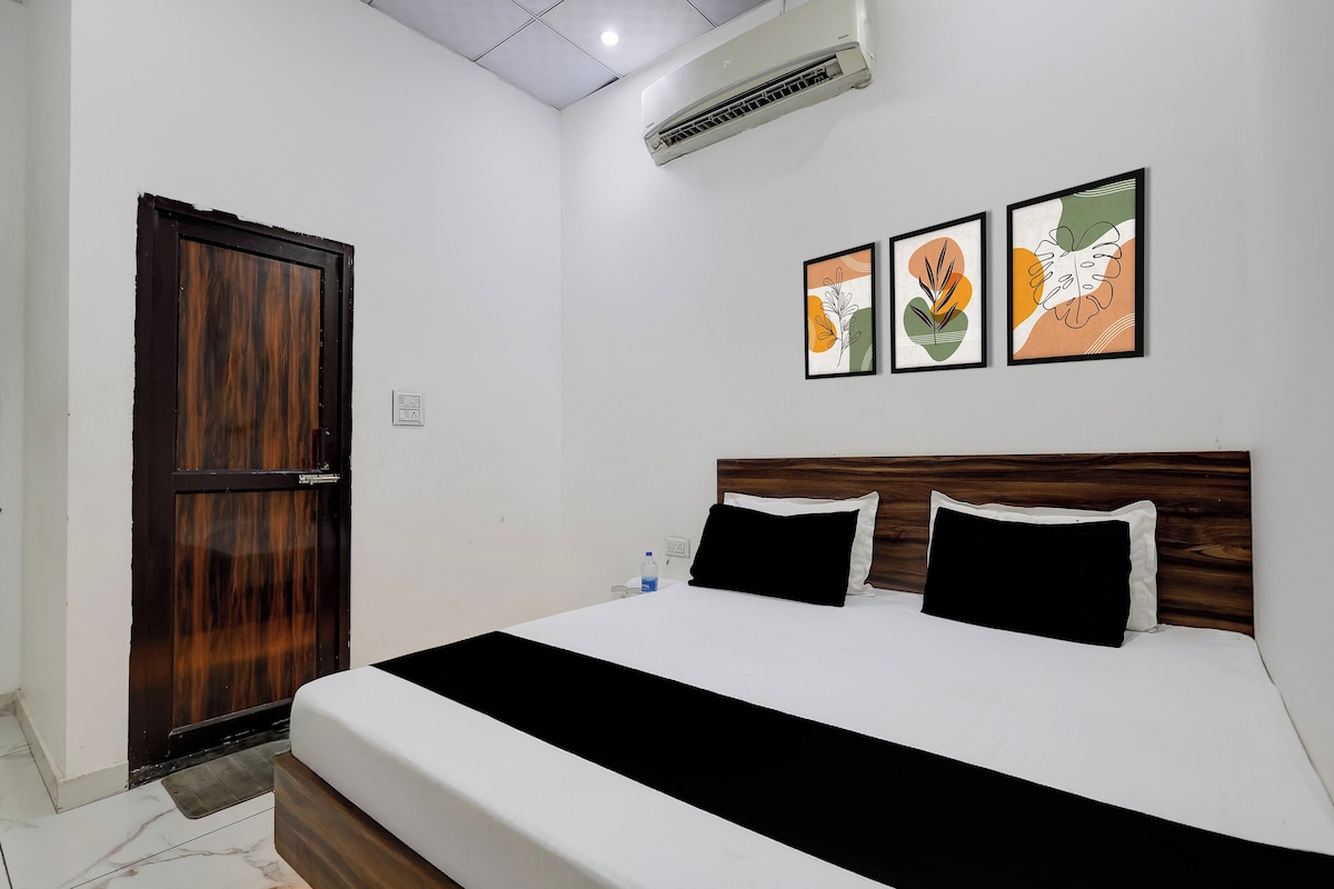 A hotel room with a wooden door, a bed with white bedding and black pillows, and two framed artworks on the wall.