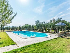 Una piscina rectangular con un borde de baldosas, un área verde con árboles y un paraguas a rayas azules.