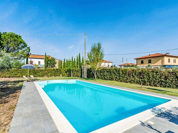 Piscina rectangular con solárium alicatado, rodeada de un seto cuidado y algunos árboles.