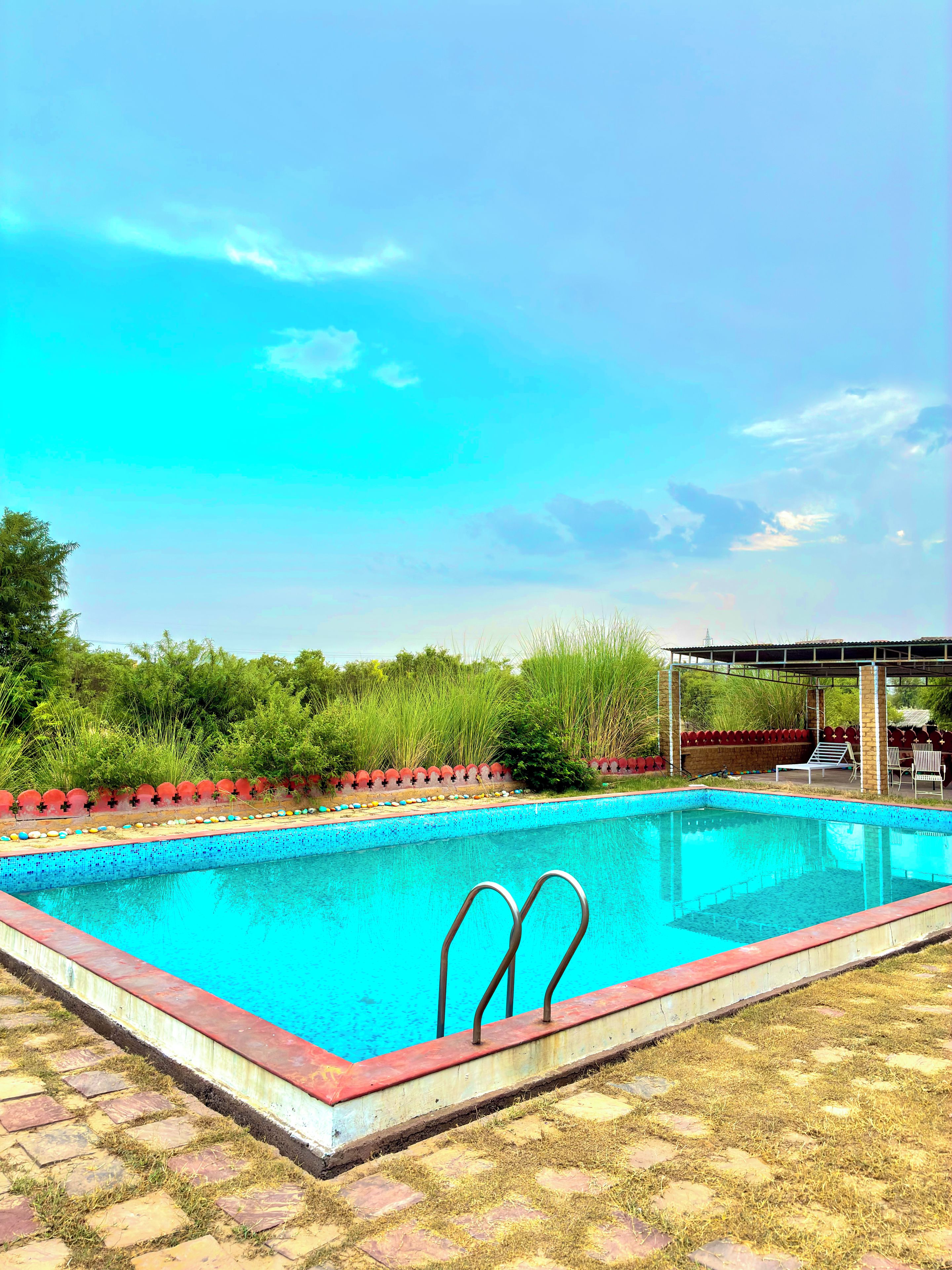 Una piscina con borde rojo, una zona de estar cubierta y un césped con valla.