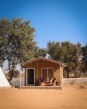 Una petita casa de maó amb un porxo, dues cadires de vimetla i una tenda a prop.