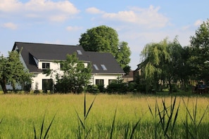 A two-story house with a sloped roof, surrounded by a grassy field and trees. - Holiday apartment with 2 bedrooms (Heidesee)