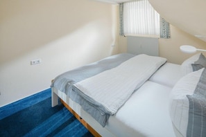 Ein Schlafzimmer mit einem Bett, einem Fenster mit Jalousien, einem Heizkörper und einem blau teppichbedeckten Treppenhaus.