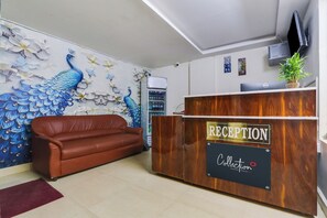 Uma área de recepção com uma mesa de madeira identificada com 'RECEPTION' e 'Collection', um sofá de couro marrom e uma parede com um mural de pavão.