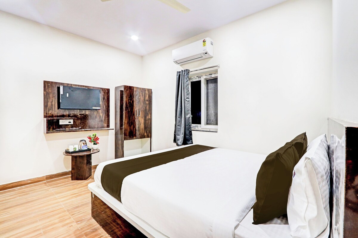 Quarto de hotel com cama, televisão na parede, janela com cortinas e uma pequena mesa com um vaso de flores.