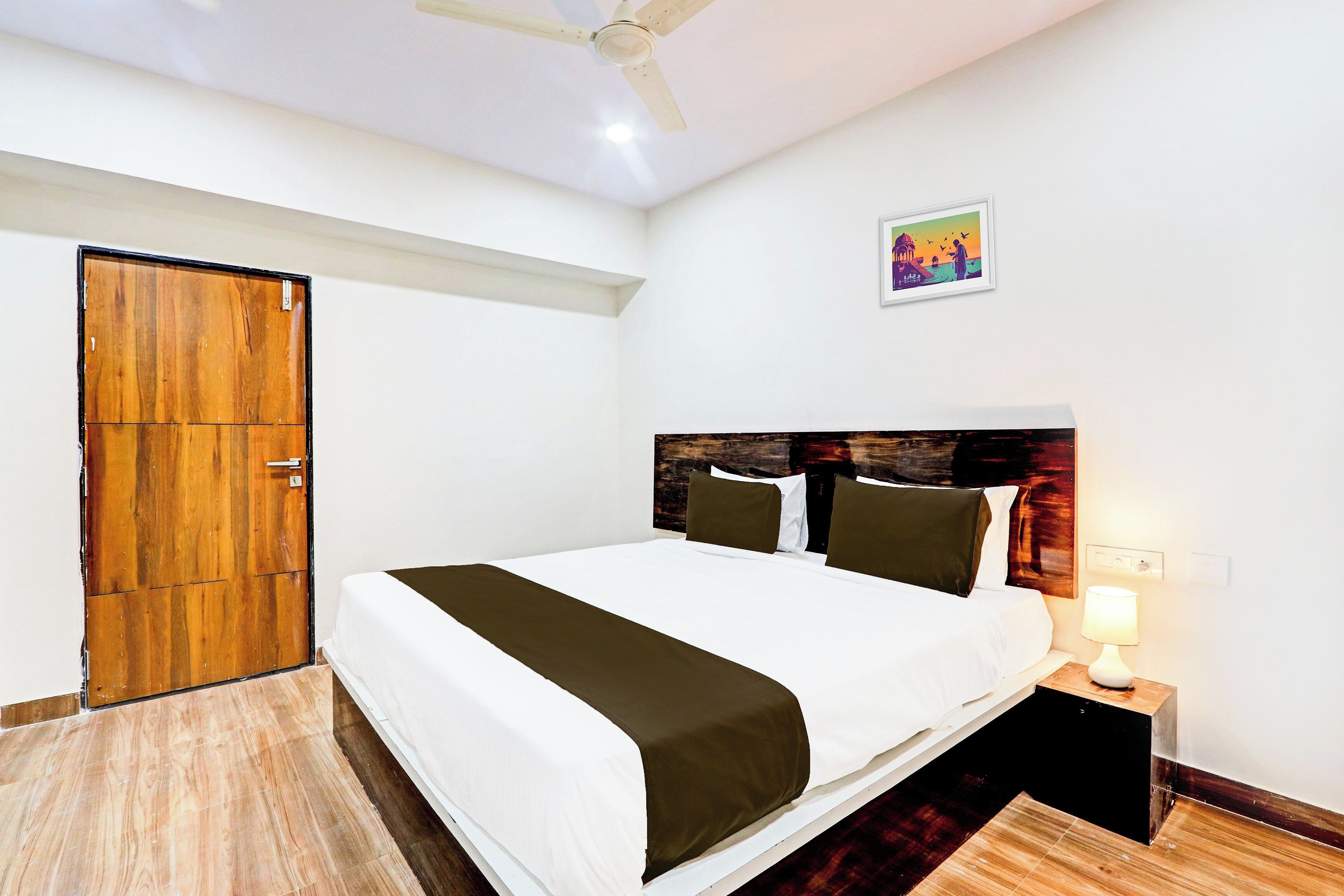 Habitación de hotel con cama, mesita de noche, lámpara de pared, ventilador de techo y puerta de madera.