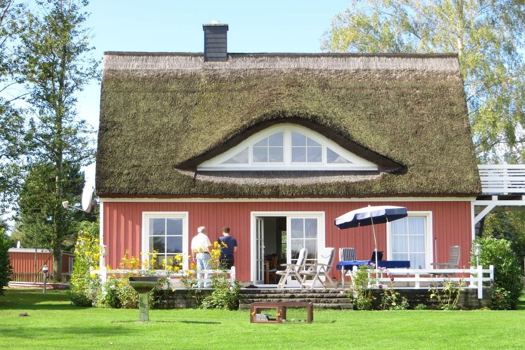 A thatched-roof house with a red exterior, a white porch, and a garden with flowers and a bench.