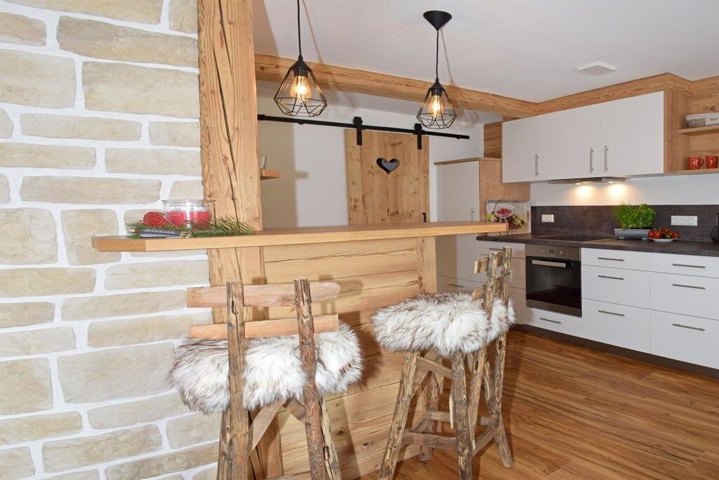 A kitchen with a stone wall, wooden bar, two bar stools with fur, and pendant lights.