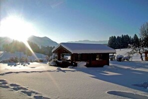 Eine Holzhütte mit schneebedecktem Dach, umgeben von verschneitem Boden und Bäumen, mit Bergen im Hintergrund.